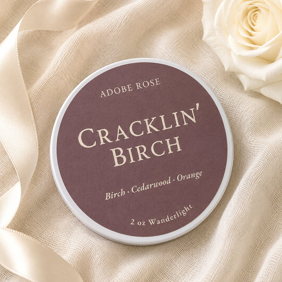 A round tin of Adobe Rose Cracklin' Birch powder paint sits on draped cream fabric with a white rose in the corner. The label reads 'ADOBE ROSE' and 'CRACKLIN' BIRCH' with 'Birch - Cedarwood - Orange' and '2 oz Wanderlight' below.