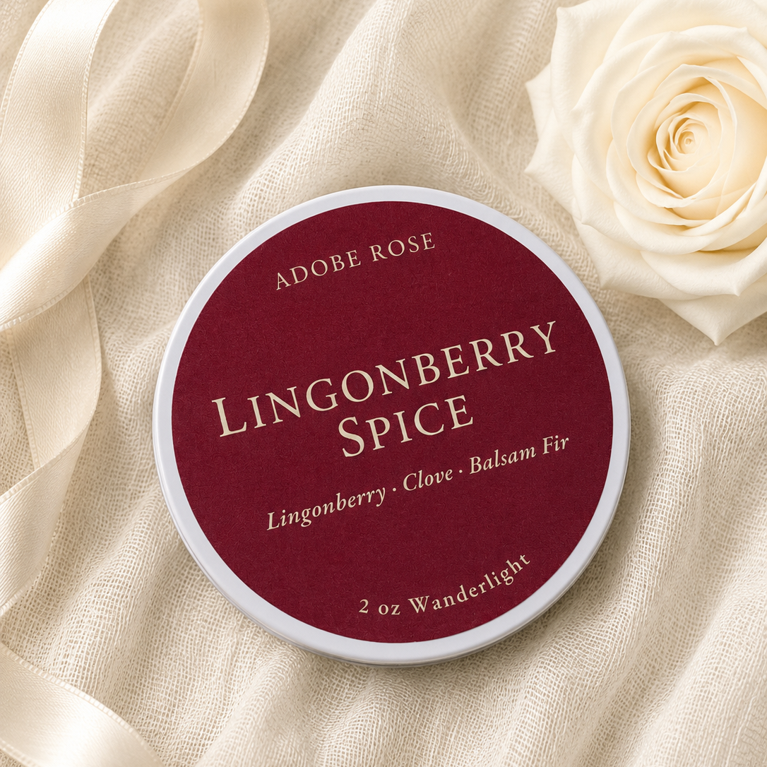 A round tin of Adobe Rose Lingonberry Spice fragrance sits on cream-colored fabric with a cream satin ribbon and a cream rose. The label is deep burgundy with cream lettering.