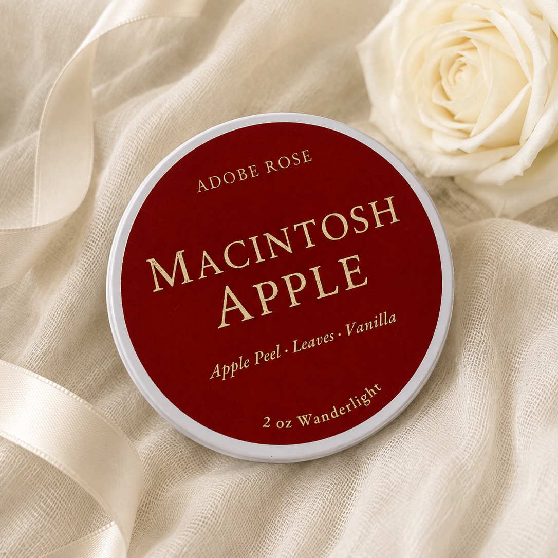 A round tin of Adobe Rose Macintosh Apple scented wax melts sits on draped cream fabric next to a white rose. The label is dark red with cream lettering.