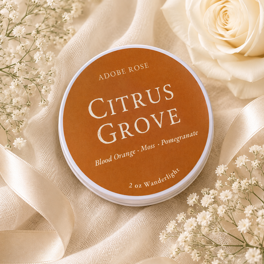 A round tin of Adobe Rose Citrus Grove fragrance oil, styled on a soft beige fabric background with white roses and baby's breath flowers.