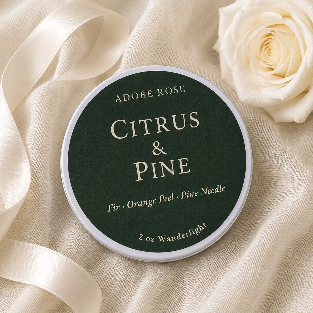 A round tin of Adobe Rose Citrus & Pine needles for fir and orange peel, styled on draped cream linen fabric next to a white rose and a cream satin ribbon.