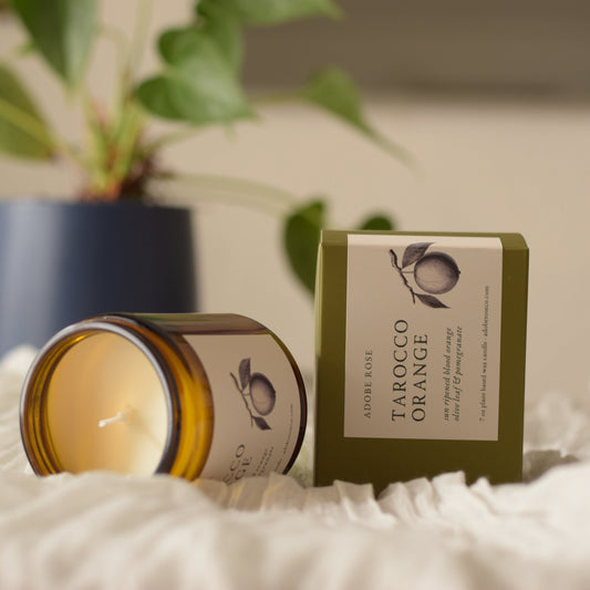 Candle and packaging labeled 'Tarocco Orange' on a soft surface with a plant in the background