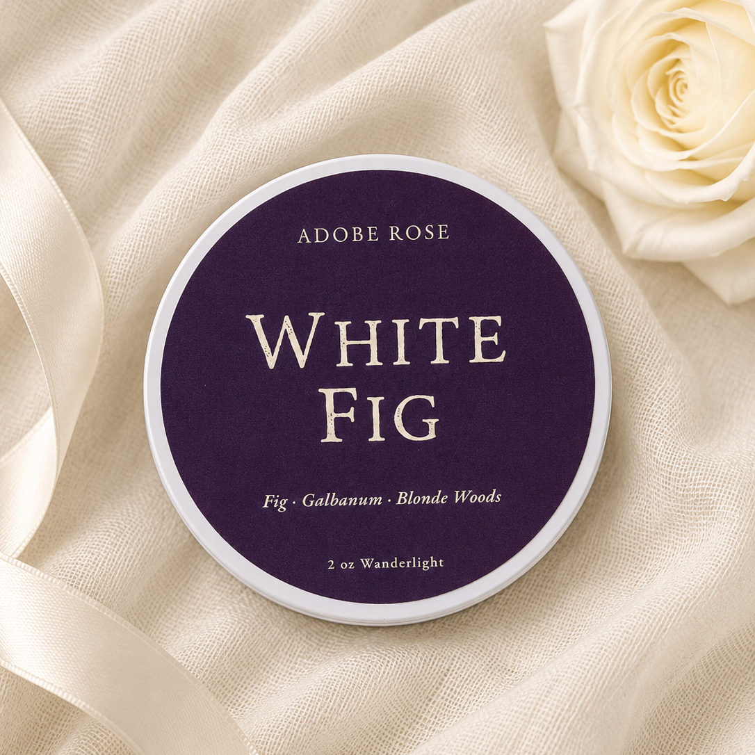 A round tin of Adobe Rose White Fig nail powder sits on draped cream fabric next to a white rose. The lid is dark purple with cream text reading 'ADOBE ROSE' and 'WHITE FIG'.