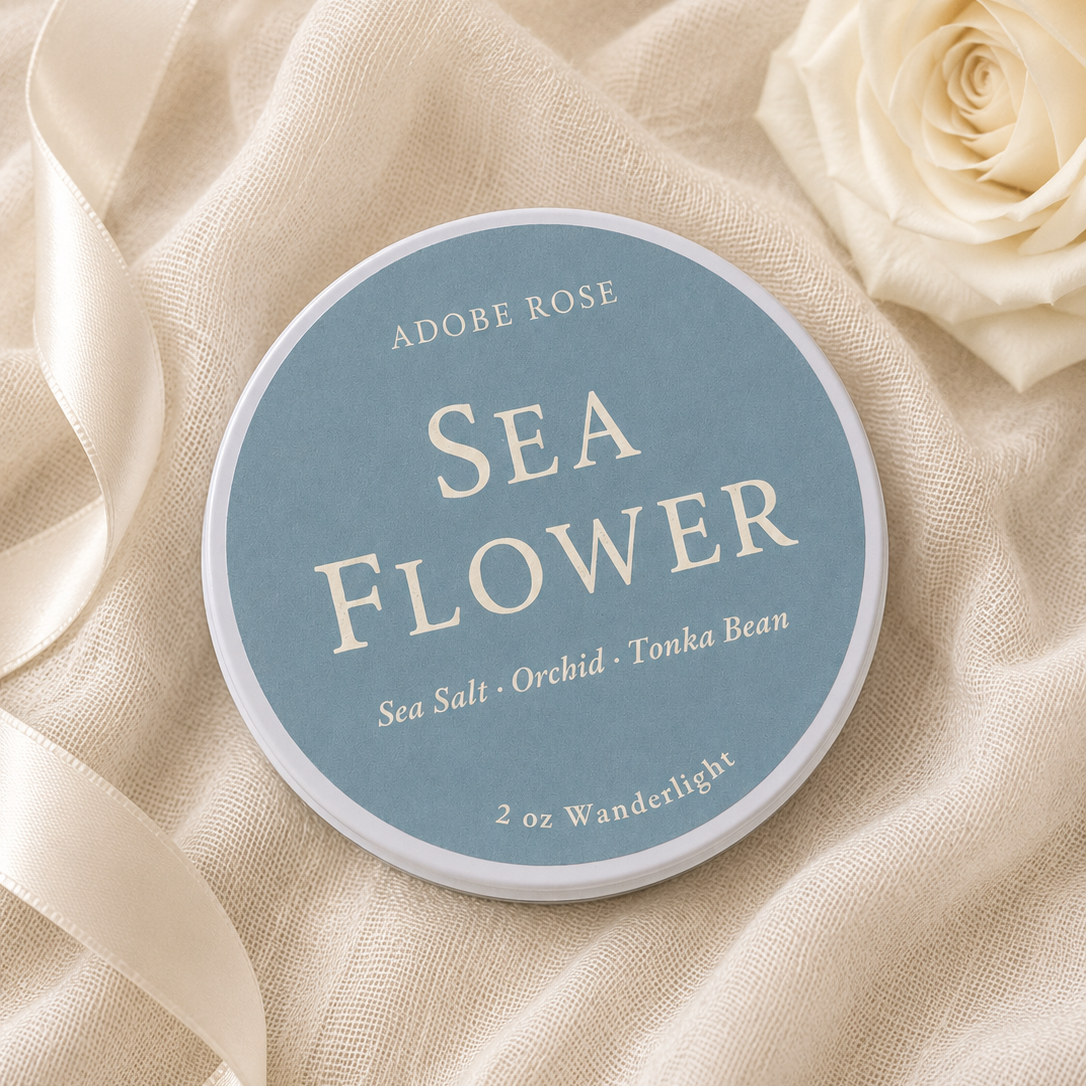 A round tin of Adobe Rose Sea Flower salt sits on draped cream fabric with a cream satin ribbon and a cream rose. The label is light blue with cream text.
