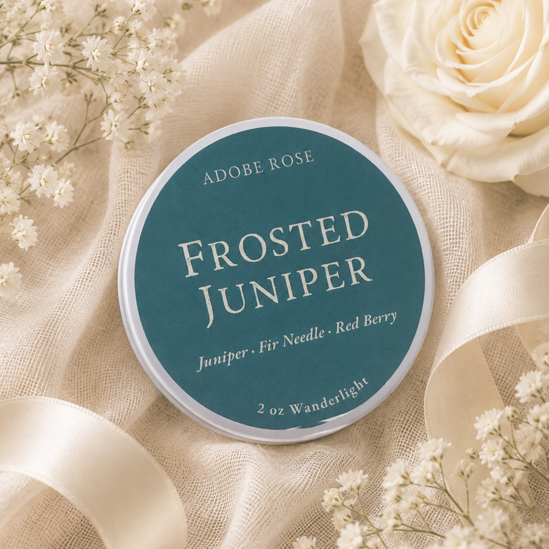 A round tin of Adobe Rose 'Frosted Juniper' craft pigment sits on draped cream fabric surrounded by white roses, baby's breath, and a cream ribbon. The tin label is teal with white text.