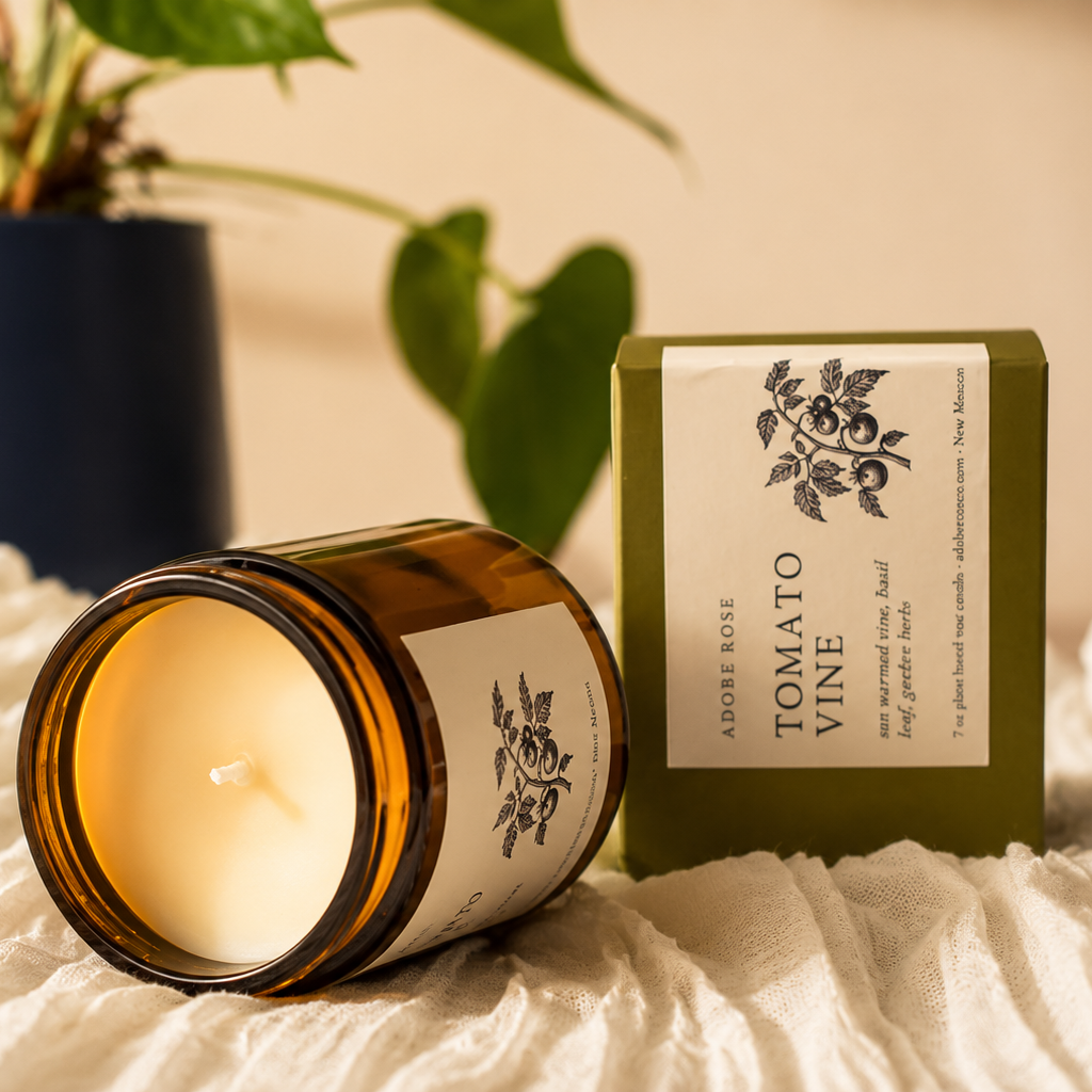 Candle in a glass jar with a decorative box labeled 'Tomato Vine' on a textured surface with a plant in the background.
