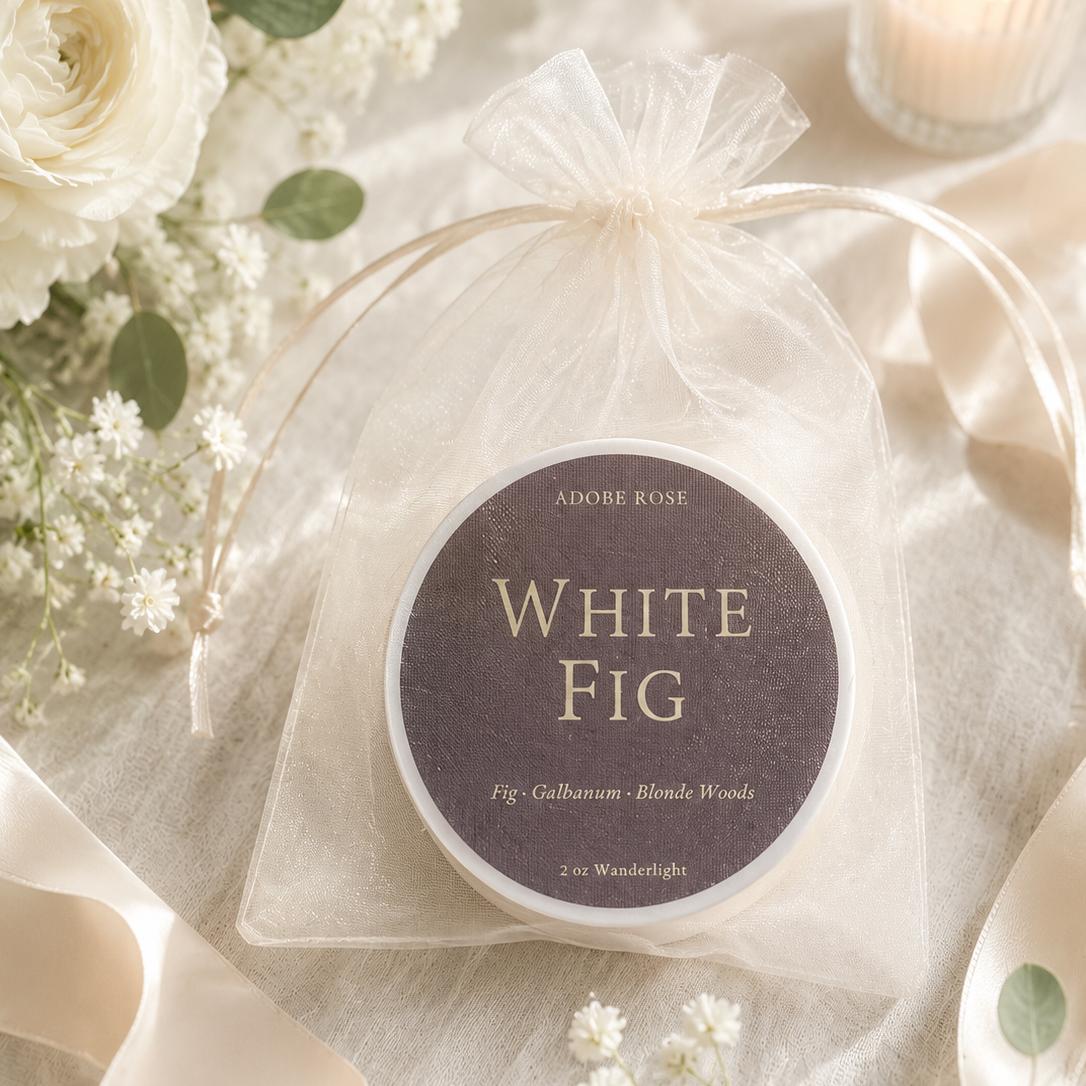 A round tin of Adore Rose White Fig wood scent oil in a sheer white drawstring pouch, styled with white roses, eucalyptus leaves, and a lit candle on a cream fabric background.