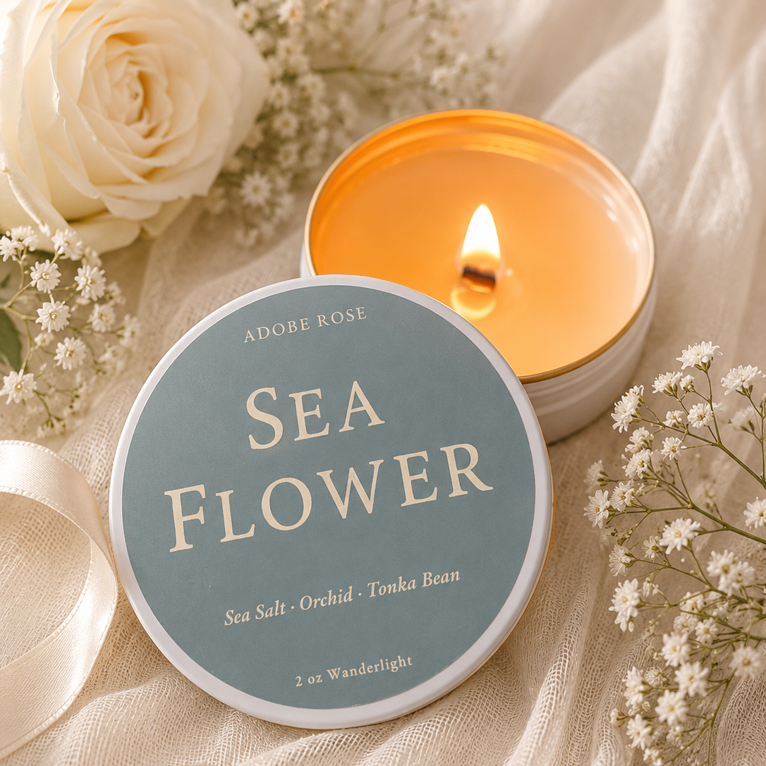 A round tin candle labeled 'ADOBE ROSE SEA FLOWER' sits on draped cream fabric beside an open flame candle and white flowers. The lid is pale blue with cream lettering, and the open candle wax is warm amber-orange.